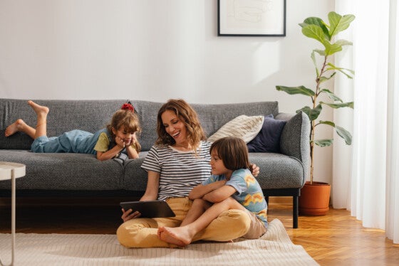 Woman with her kids sitting on the floor and using digital tablet for playing games, watching movies and relaxing at home. Her son is cuddled with her, while the daughter is lying on the sofa next to them. They are all smiling and looking down at the screen.