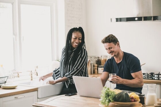 Happy married interracial couple looking at laptop computer together while doing daily moring routines in the kitchen at home