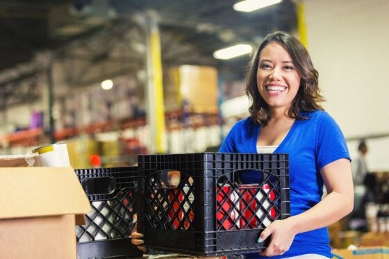 Young adult Asian and Hispanic American woman is smiling and looking at camera. She is holding a crate of donated grocery and food items. She's volunteering in a large food bank warehouse. Woman is sorting donated food to distribute to community.