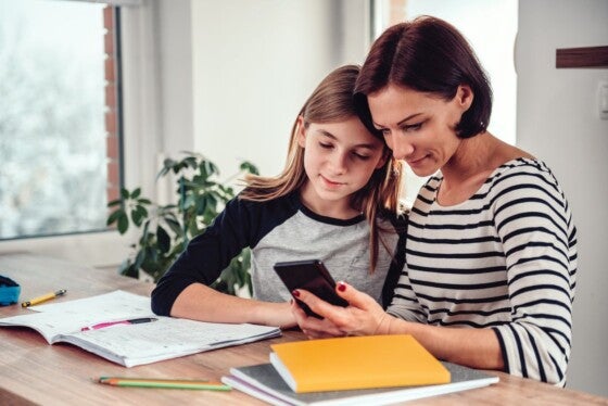 Mother using smart phone and helping daughter with homework at the table in dining room by the window