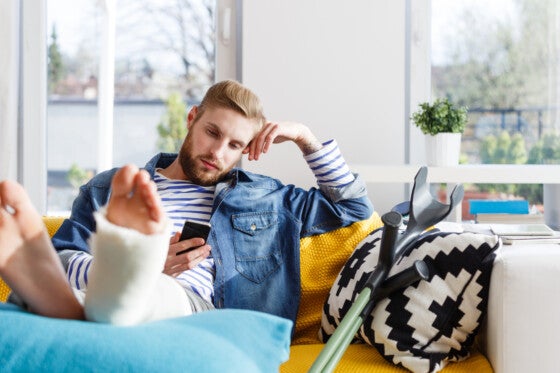 Young man with broken leg in plaster cast lying down on sofa at home and using a smart phone.