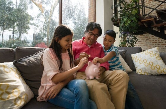 Grandfather teaching his grandkids to save money in piggy bank while sitting on couch at home all smiling