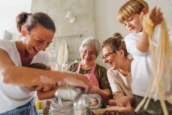 Photo of a smiling senior woman teaching her daughters and grandsons how to make homemade pasta in the kitchen; having a great time while making food and enjoying each other's company.